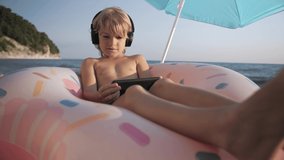 Child video chats on smart phone on beach on blue water background. Boy with headphones listen to music and singing, using portable handheld device on summer vacation. Doing selfie. Wide angle view