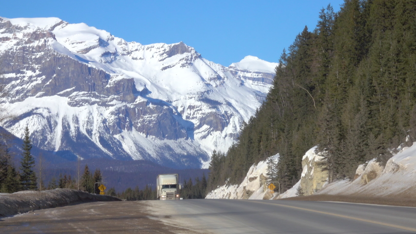 Semi-trailer truck and car drive along the highway crossing Jasper National Park offering a spectacular view of a snow capped mountain range and lush spruce forests. Lorry hauls cargo across Alberta.