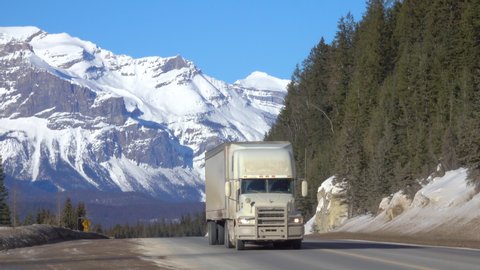 Semi-trailer truck and car drive along the highway crossing Jasper National Park offering a spectacular view of a snow capped mountain range and lush spruce forests. Lorry hauls cargo across Alberta.