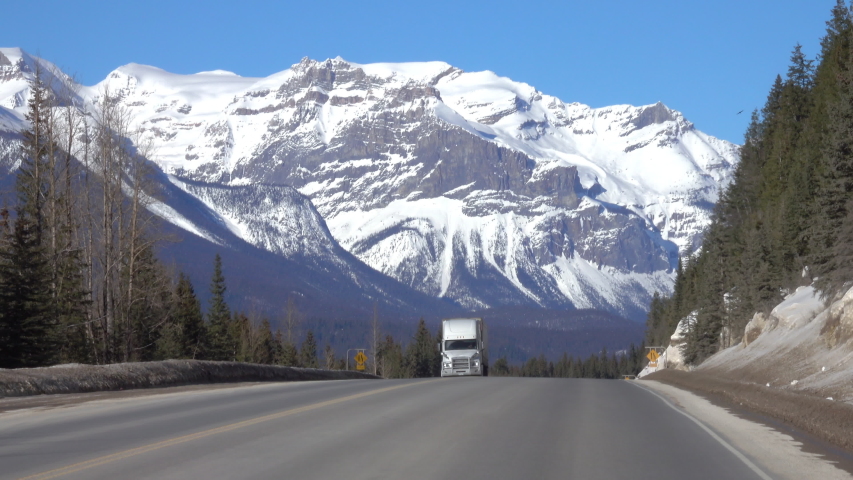 Three trucks haul cargo down the highway leading towards the breathtaking snow capped mountains in Jasper National Park. Lorries travel along the famous Icefields Parkway on a sunny winter day. Royalty-Free Stock Footage #1044285670