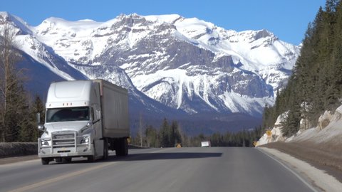 Three trucks haul cargo down the highway leading towards the breathtaking snow capped mountains in Jasper National Park. Lorries travel along the famous Icefields Parkway on a sunny winter day.