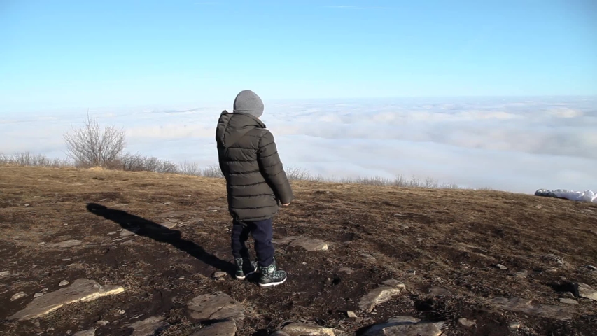 Little boy in coat and hat standing on top of mountain and looks into distance at clouds