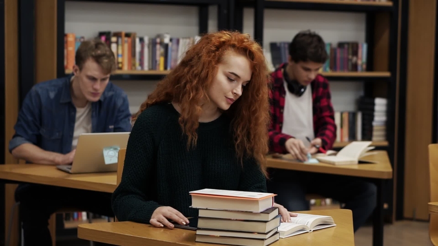 Portrait of a young red headed curly female student working on her thesis or preparing for exam. Reading a book and typing important on tablet sitting at the table in library. Male classmates on