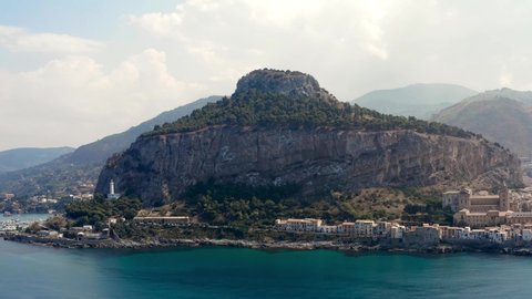 Aerial view of breathtaking mountain located at the sea shore and coastal town. Stock footage. Flying over the turquoise water surface and the stony shore on blue cloudy sky background.