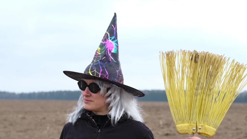 Outdoor portrait of funny woman in witch costume with cone hat, grey wig and modern broom smiling or grinning at camera. Halloween concept.