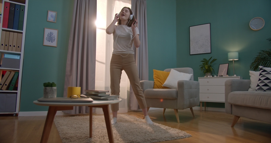 Pretty Caucasian young girl in headphones listening to the music on the smartphone, dancing and singing in the cozy living room. Indoors. Stay at home. Royalty-Free Stock Footage #1044345970