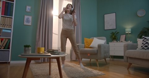 Pretty Caucasian young girl in headphones listening to the music on the smartphone, dancing and singing in the cozy living room. Indoors. Stay at home.