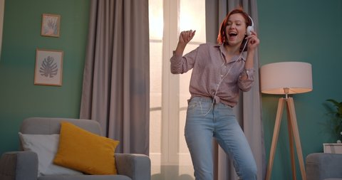 Red-haired young Caucasian beautiful woman dancing happily in the living room in her apartment with headphones on her head. Stay home