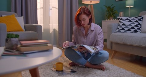 Young stylish charming red-haired woman reading a fashion magazine o the floor in the cozy living room on weekend. Inside. Quarantine, virus, stay at home