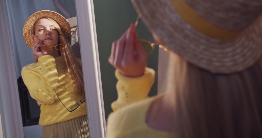View over the shoulder on the Caucasian young pretty blonde girl fashion blogger in a hat looking in the mirror while trying on sunglasses. Indoors.