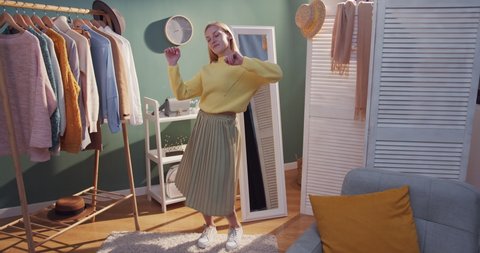 Blonde young Caucasian attractive girl with long hair in skirt and yellow sweater trying on clothes in the cozy big wardrobe and dancing. Indoors.