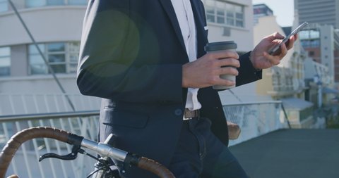 Side view mid section of a young Caucasian businessman on the go in the city, using smartphone and holding a cup of takeaway coffee in the street leaning on a bike with buildings in the background,