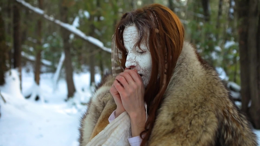 Close up footage of a native spiritual elder with traditional fur skin coat, a cracked white face and eagle feather, blowing on cold hands in winter