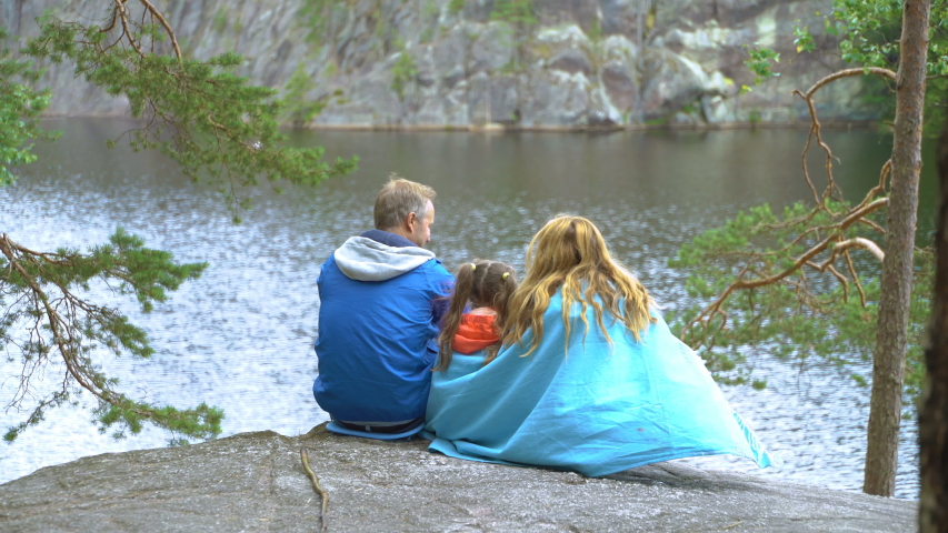 Family with child sitting on the rocky shore of a forest lake in Finland wrapped in a blanket