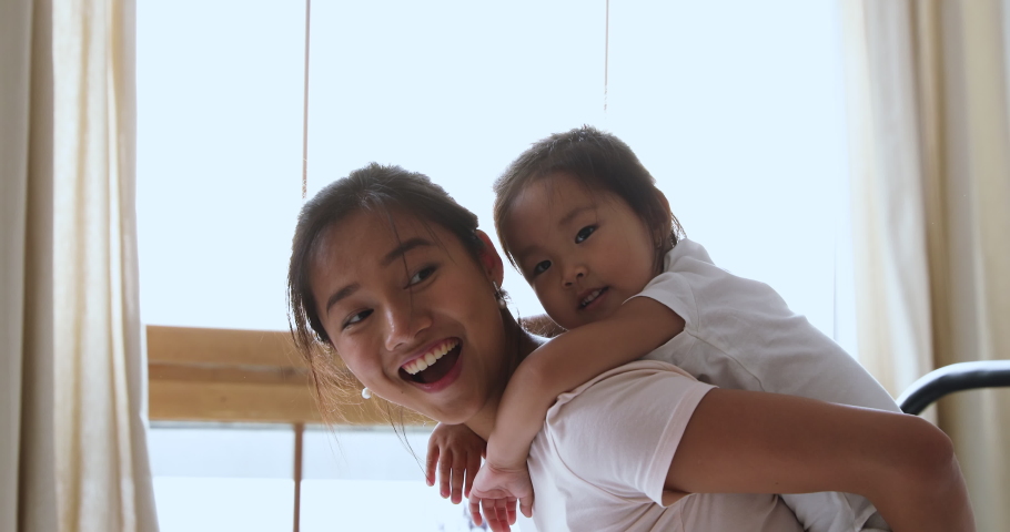 Smiling young adult asian mum piggybacking little cute child daughter hugging mother from her back playing laughing in bed, small adorable vietnamese kid girl embracing mommy having fun in morning Royalty-Free Stock Footage #1044388174