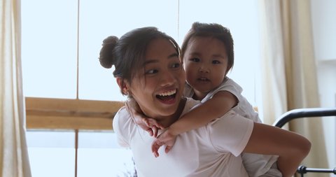 Smiling young adult asian mum piggybacking little cute child daughter hugging mother from her back playing laughing in bed, small adorable vietnamese kid girl embracing mommy having fun in morning 스톡 비디오
