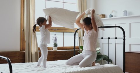 Funny happy asian small cute kid daughter and parent mum holding pillows playing fight in bed, little vietnamese child girl having fun with adult mother enjoying lifestyle morning activity together
