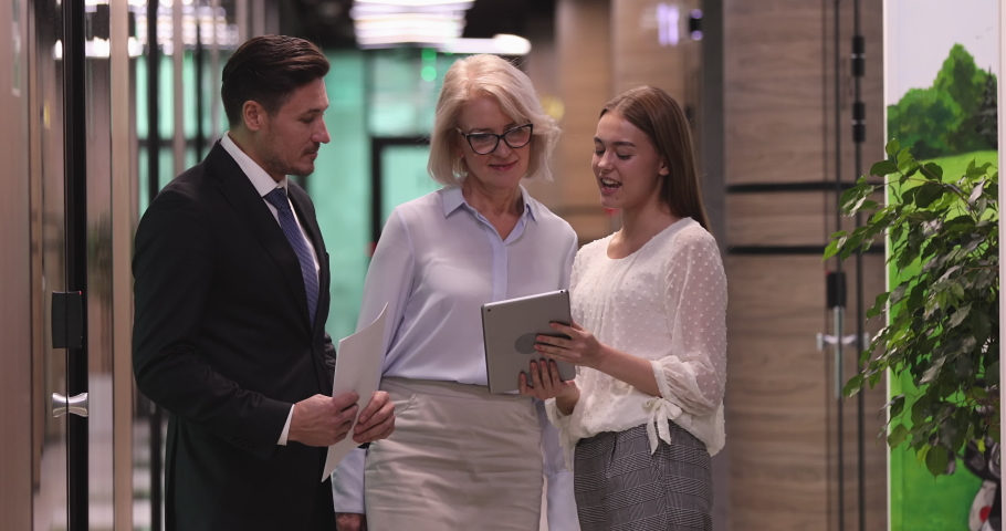 Young intern trainee meeting executive old mentor colleagues instructing employee showing plan on digital tablet walking in hallway, three friendly business team people talk stand in modern office Royalty-Free Stock Footage #1044388228