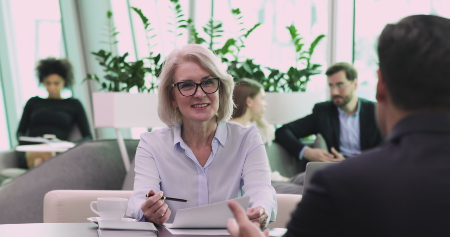 Smiling older mature business woman employer hr manager staffing holding cv checking male candidate resume asking questions during job interview office meeting, work placement and recruitment concept