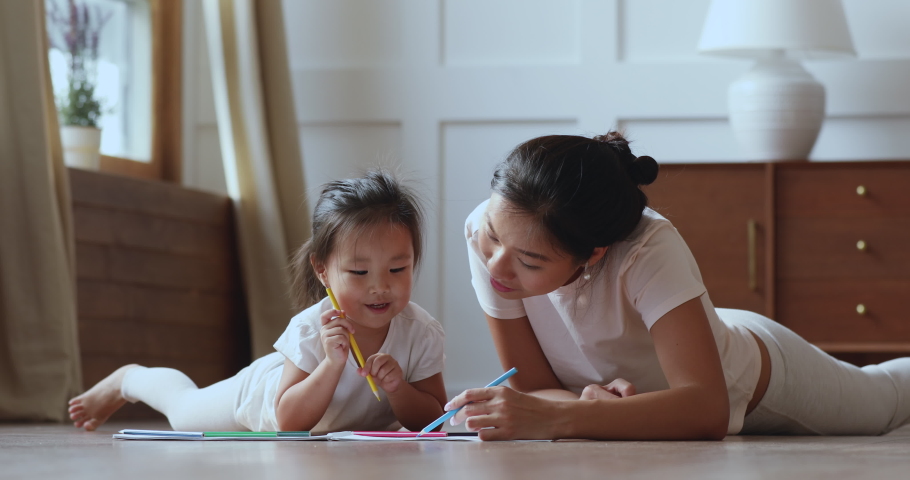 Cute adorable asian ethnic kid girl holding color pencil learn drawing lying on warm floor with young vietnamese mom daycare babysitter talking playing teaching helping little child daughter at home Royalty-Free Stock Footage #1044388309