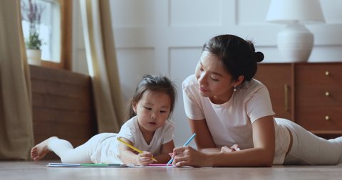 Cute adorable asian ethnic kid girl holding color pencil learn drawing lying on warm floor with young vietnamese mom daycare babysitter talking playing teaching helping little child daughter at home