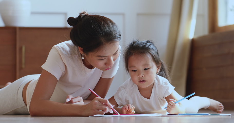 Caring young asian ethnic mum or nanny helping cute kid daughter teaching toddler child daughter drawing picture with pencils lying on warm heated floor in bedroom learn creative art activity at home Royalty-Free Stock Footage #1044388318