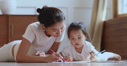 Caring young asian ethnic mum or nanny helping cute kid daughter teaching toddler child daughter drawing picture with pencils lying on warm heated floor in bedroom learn creative art activity at home