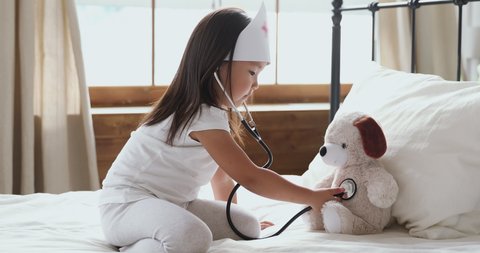 Cute little asian child girl wear medical hat playing game as vet doctor holding stethoscope listening sick dog in bed, adorable preschool korean child pretending doctor treat toy patient in bedroom