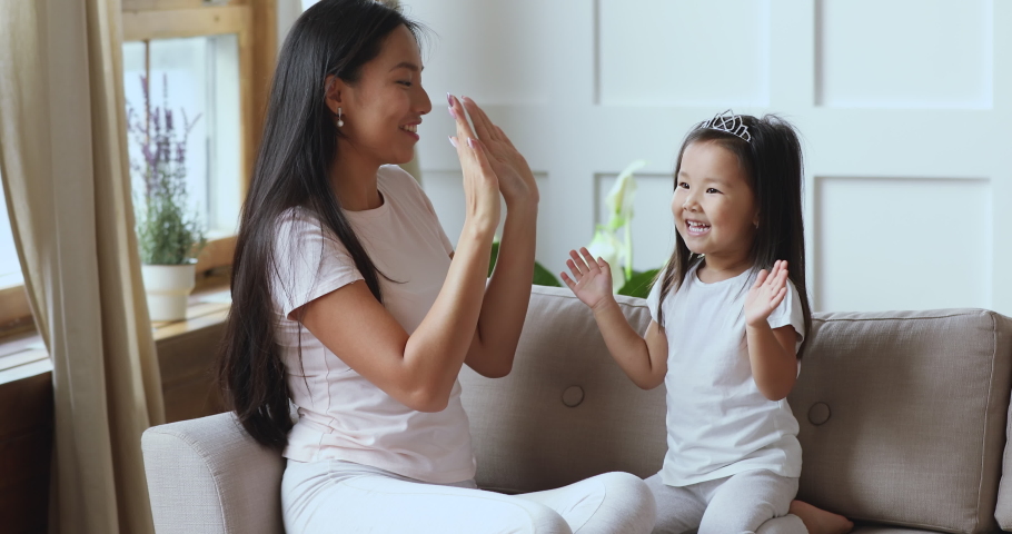 Happy asian mum and cute toddler child daughter wear crowns having fun enjoy leisure game playing patty cake at home, smiling young mother and little kid girl clap hands laughing relaxing on sofa Royalty-Free Stock Footage #1044395572