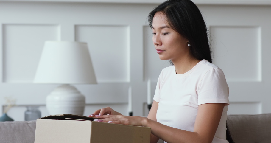 Happy amazed young asian girl consumer holding opening cardboard box sit on sofa at home, excited vietnamese woman customer receive unbox good parcel, order postal shipping courier delivery concept | Shutterstock HD Video #1044395584