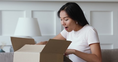 Happy amazed young asian girl consumer holding opening cardboard box sit on sofa at home, excited vietnamese woman customer receive unbox good parcel, order postal shipping courier delivery concept