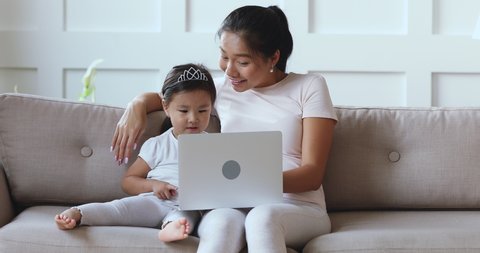 Happy funny cute little asian kid daughter learning using technology device look at laptop enjoy watching online cartoons on computer with adult parent vietnamese mom or nanny resting on sofa at home