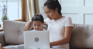 Caring asian mother teaching cute adorable small kid daughter using laptop online app for children education entertainment, happy little child girl learning computer tech sit on sofa with parent mum