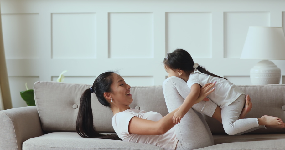 Happy young asian mommy playing with cute small adorable toddler child daughter wear crown holding lifting funny little vietnamese kid up having fun fly in mum arms doing gymnastics exercise on sofa Royalty-Free Stock Footage #1044395617
