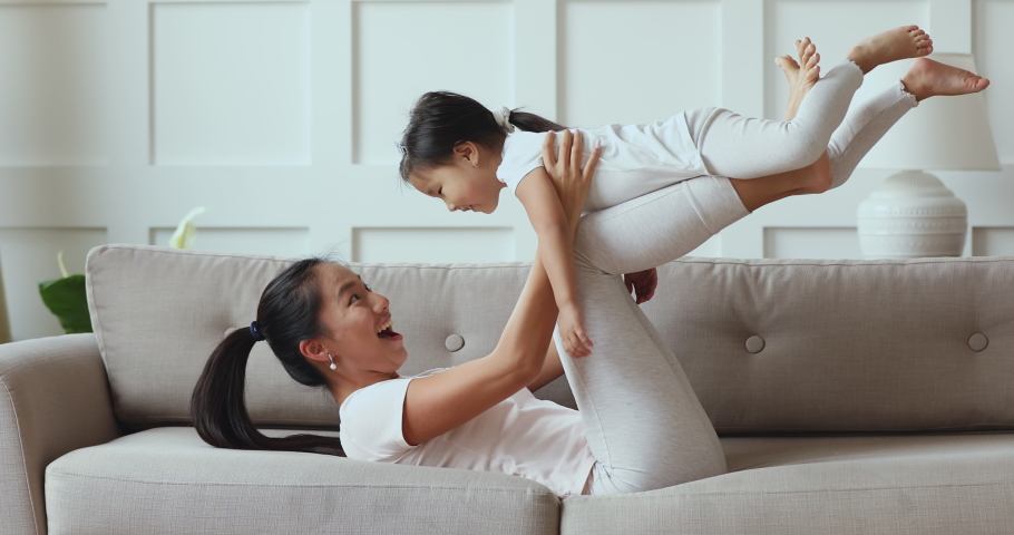 Happy young asian mommy playing with cute small adorable toddler child daughter wear crown holding lifting funny little vietnamese kid up having fun fly in mum arms doing gymnastics exercise on sofa | Shutterstock HD Video #1044395617