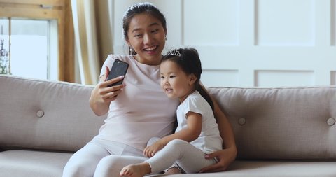 Happy family young asian mom and cute little kid daughter wear crown using smart phone funny ar app recording social media videos or taking selfie having fun enjoying tech gadget sit on sofa at home