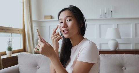 Millennial asian ethnic girl video blogger influencer holding cosmetic product concealer looking at camera recording beauty blog at home shooting social media makeup tutorial vlog channel concept