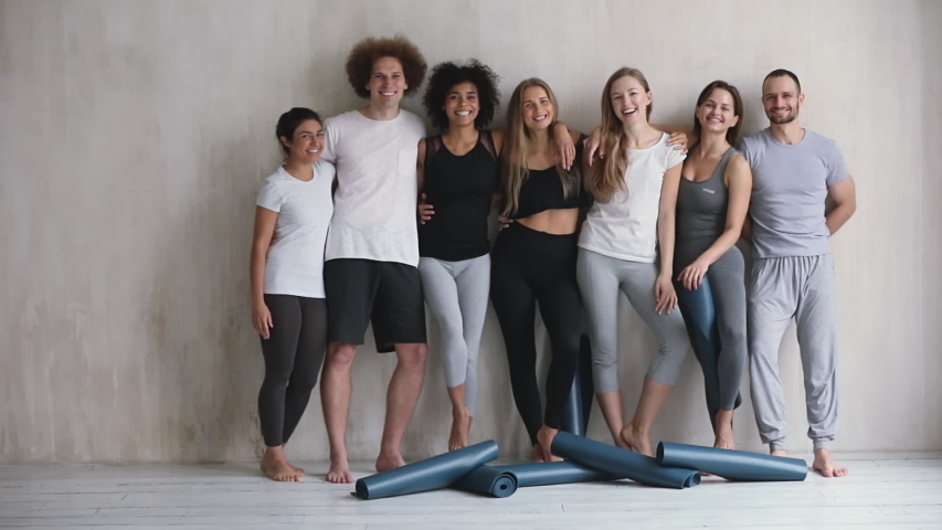 Full length view seven multi-ethnic cheery young friends girls and guys embracing standing leaned on wall waiting for yoga class, finished work out feels happy, on floor lot of mats, wellness concept Royalty-Free Stock Footage #1044404398