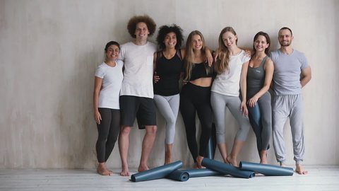 Full length view seven multi-ethnic cheery young friends girls and guys embracing standing leaned on wall waiting for yoga class, finished work out feels happy, on floor lot of mats, wellness concept