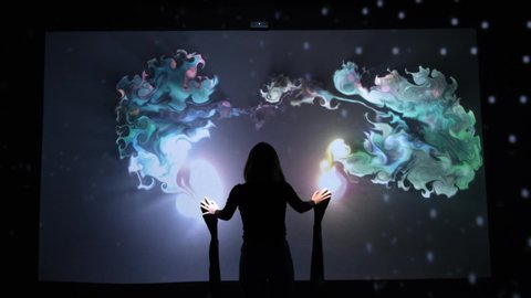 Girl plays with interactive video installation, hands draw the shape heart. New art form, generative graphics. Silhouette of girl draws multi-colored paints interactive installation.