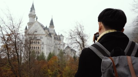 Back view of male Asian tourist in black jacket with backpack and camera and taking photos of view of Neuschwanstein castle in haze autumn forest during trip to Bavaria, Germany
