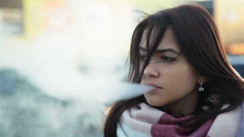 Pensive woman vaping e-cigarette between  cars on the parking zone near the mall, close up