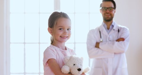 Caring male doctor pediatrician wear white uniform hugging little cute child girl patient hold toy look at camera, children healthcare professional medic treatment and protection concept, portrait