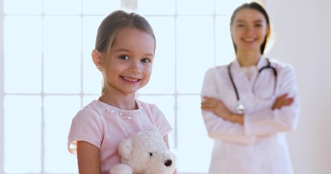 Female professional doctor pediatrician wear white coat come to happy cute little child girl embrace kid patient hold toy look at camera, children healthcare and paediatric medical treatment concept