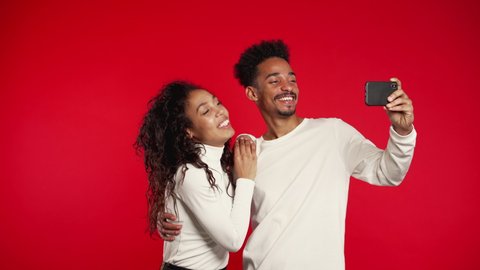 Young african american couple in white making selfie with smartphone on red studio background. Love, holidays, happiness concept.