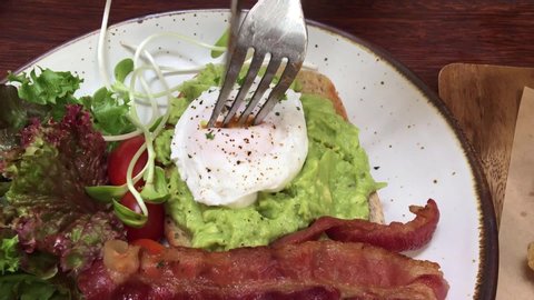 Cutting perfect poached eggs with avocado paste on toast ,crispy fried bacon ,fresh green salad that calls Open Faced Avocado Spread Sandwiches recipe in restaurant as healthy breakfast ,bunch menu