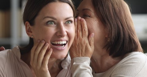 Funny two generation women family friends share secrets gossip having fun together, older mature mother whisper news rumors in ear of excited young adult daughter laugh bonding at home, closeup view