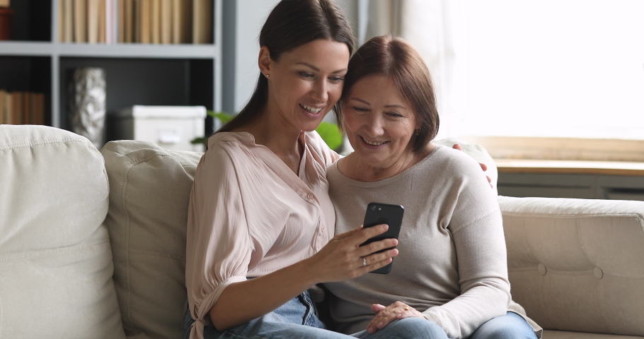 Happy two generation women family laugh look at cellphone, cheerful older mature mom and young adult daughter having fun use smart phone take selfie watch funny social media video at home sit on sofa Royalty-Free Stock Footage #1044454915