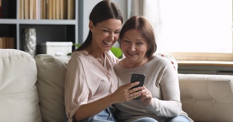 Happy two generation women family laugh look at cellphone, cheerful older mature mom and young adult daughter having fun use smart phone take selfie watch funny social media video at home sit on sofa