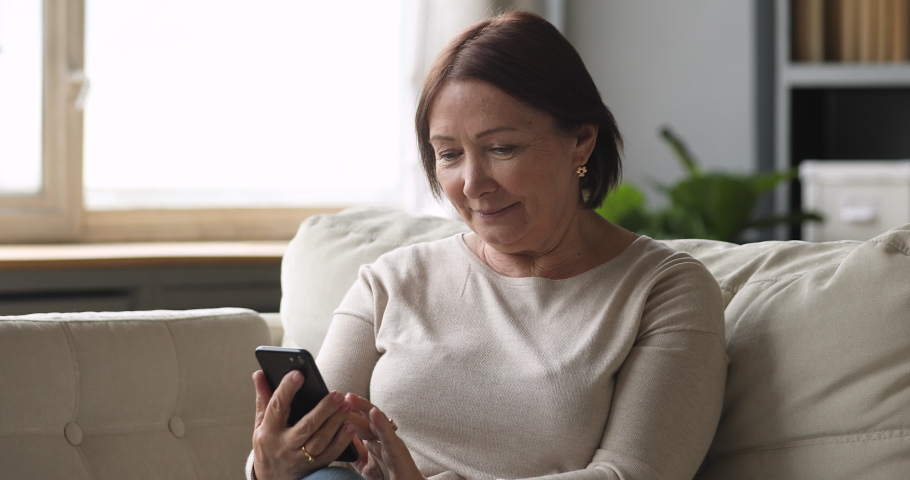 Smiling senior lady using smartphone texting message sit on couch, middle aged woman grandma hold phone typing sms enjoying communication in mobile app at home, older people and modern tech concept | Shutterstock HD Video #1044454930